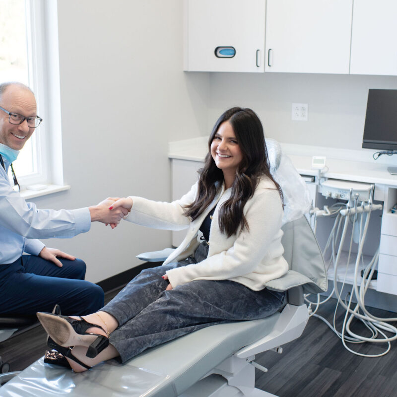 Dentist shaking hands with a smiling female patient seated in a dental chair inside a modern dental office, with dental equipment visible nearby.